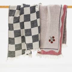 Two woven kitchen towels hanging from a wooden rod against a white background. The left blanket has a black and off-white checkerboard pattern with white fringe.
