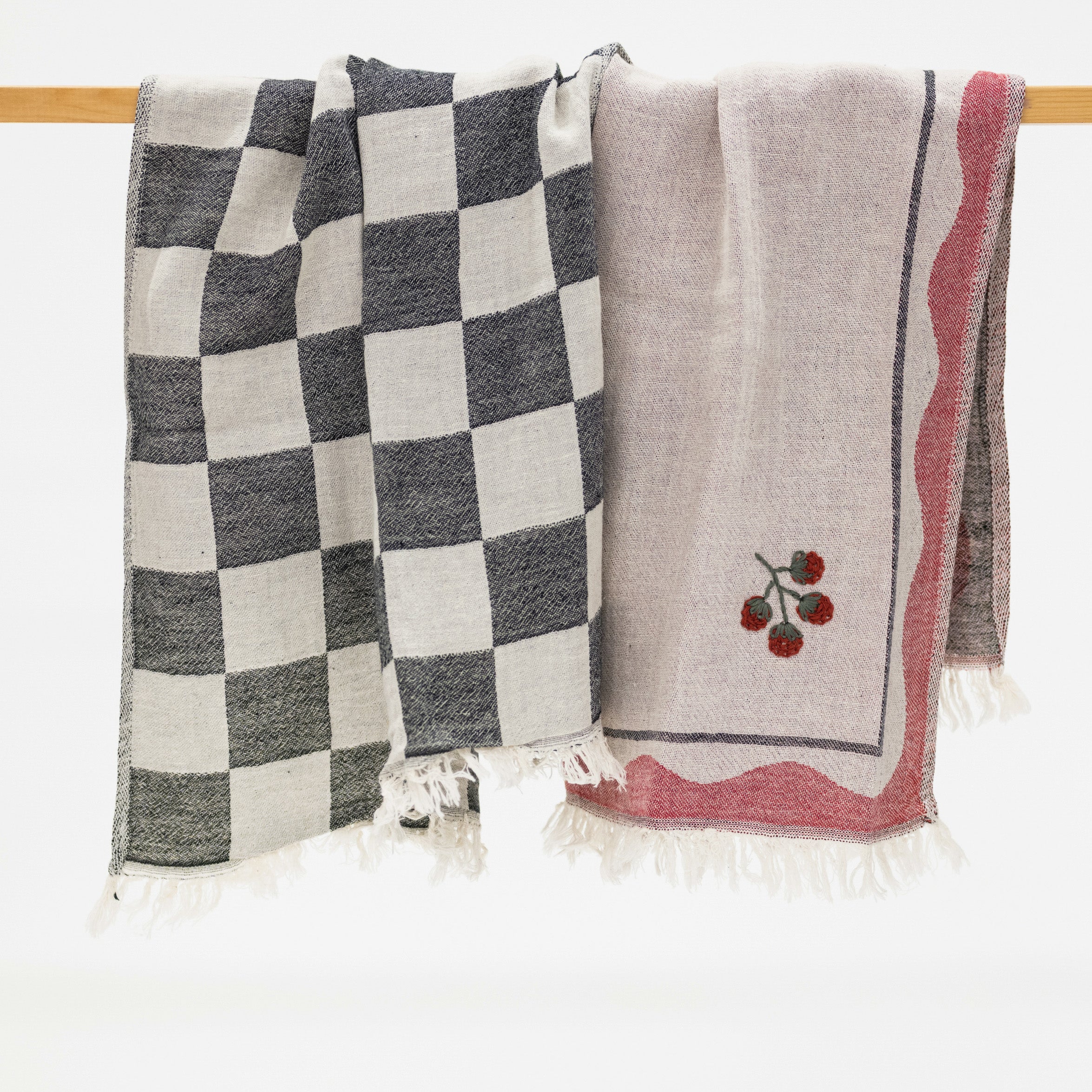 Two woven kitchen towels hanging from a wooden rod against a white background. The left blanket has a black and off-white checkerboard pattern with white fringe.