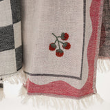 A close-up of a kitchen towel in light beige with a pink border, white fringe, and an embroidered red tomato motif on a white background.