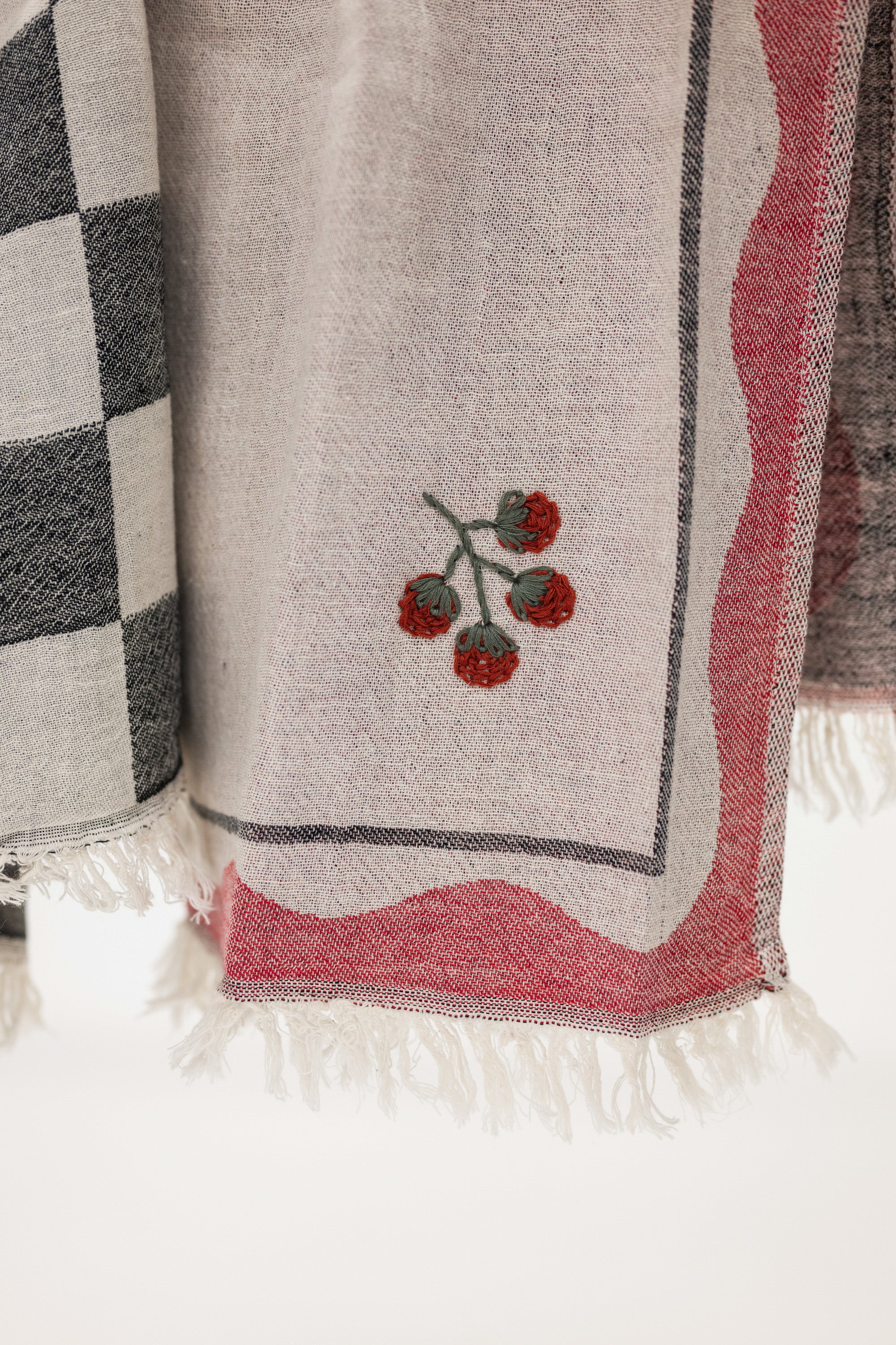 A close-up of a kitchen towel in light beige with a pink border, white fringe, and an embroidered red tomato motif on a white background.