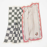 A pair of woven kitchen towels: one features a black and white checkerboard pattern, and the other has a light beige center with a pink scalloped border and a small embroidered tomato motif in the corner.