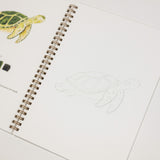 An open spiral-bound art journal or coloring book page showing a painted sea turtle on the left, a color palette strip, and a faint outline of the same turtle on the right, photographed on a white background.