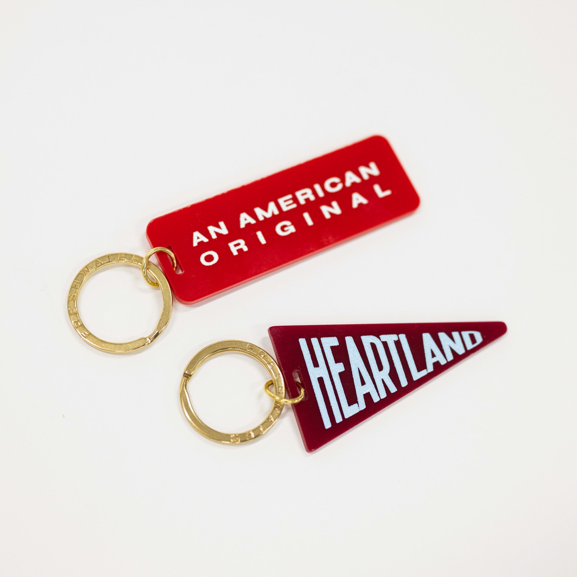 Two novelty keychains on a plain white background: a red rounded rectangle keychain with white text reading 'AN AMERICAN ORIGINAL' and a dark red triangular pennant keychain with white text reading 'HEARTLAND'. Both have gold-tone split rings.