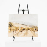 A horizontal landscape painting on canvas, displayed on a black metal easel stand against a clean white background. The painting features a soft, impressionistic brushstroke style depicting rolling hills in warm beige, tan, and brown tones under a pale sky.
