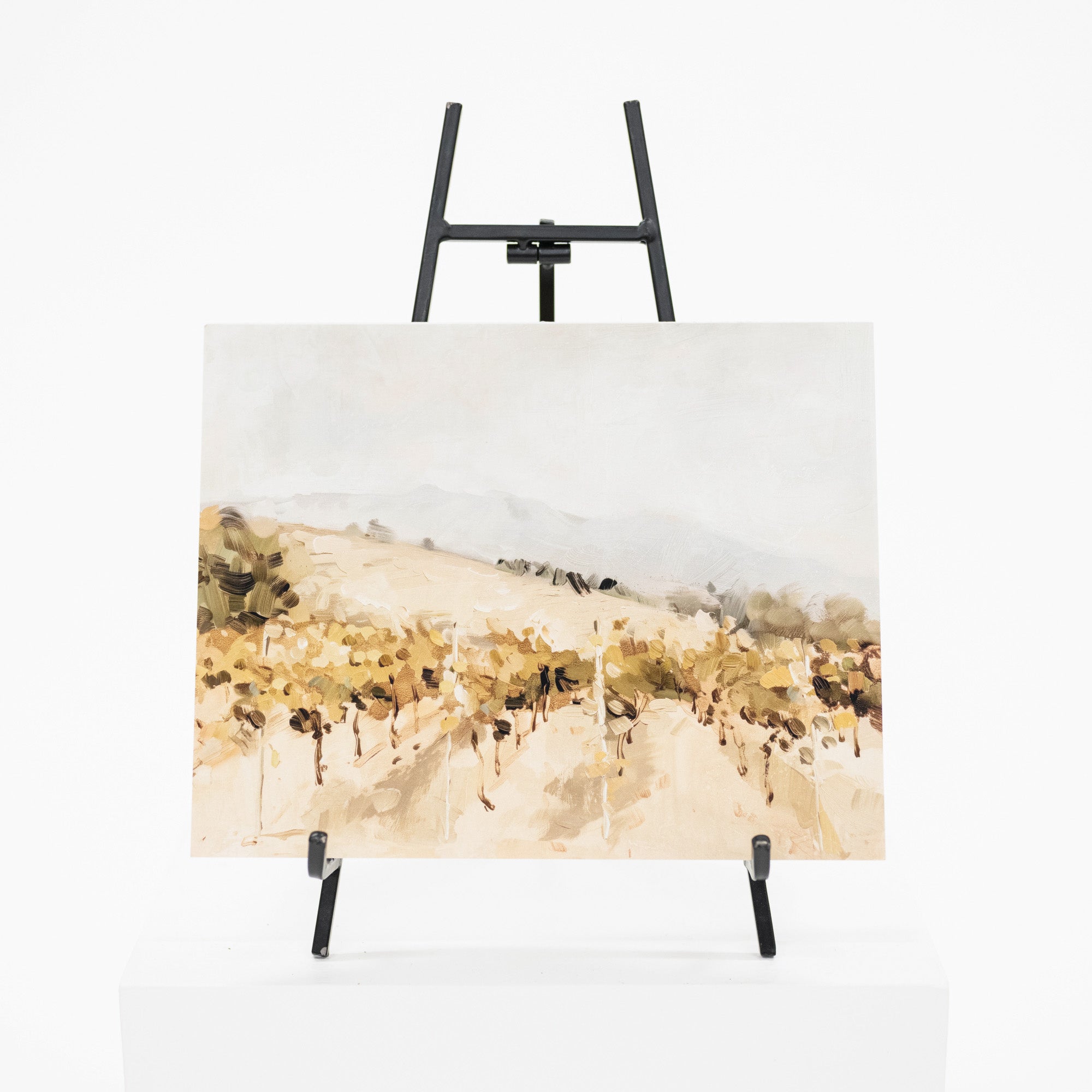 A horizontal landscape painting on canvas, displayed on a black metal easel stand against a clean white background. The painting features a soft, impressionistic brushstroke style depicting rolling hills in warm beige, tan, and brown tones under a pale sky.