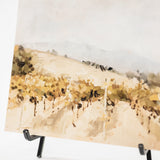 A vertical watercolor-style landscape painting of a vineyard or field with muted beige, brown, and soft blue tones, displayed on a black easel against a white background.