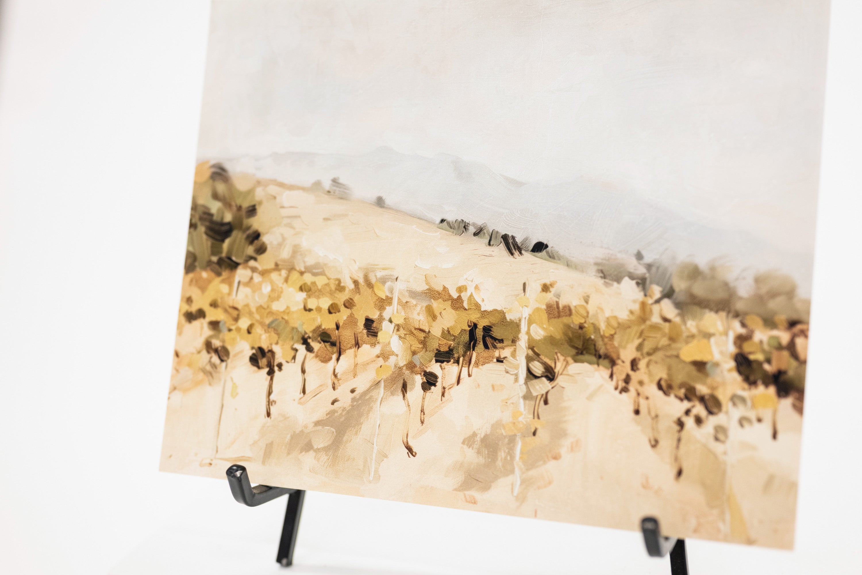 A vertical watercolor-style landscape painting of a vineyard or field with muted beige, brown, and soft blue tones, displayed on a black easel against a white background.
