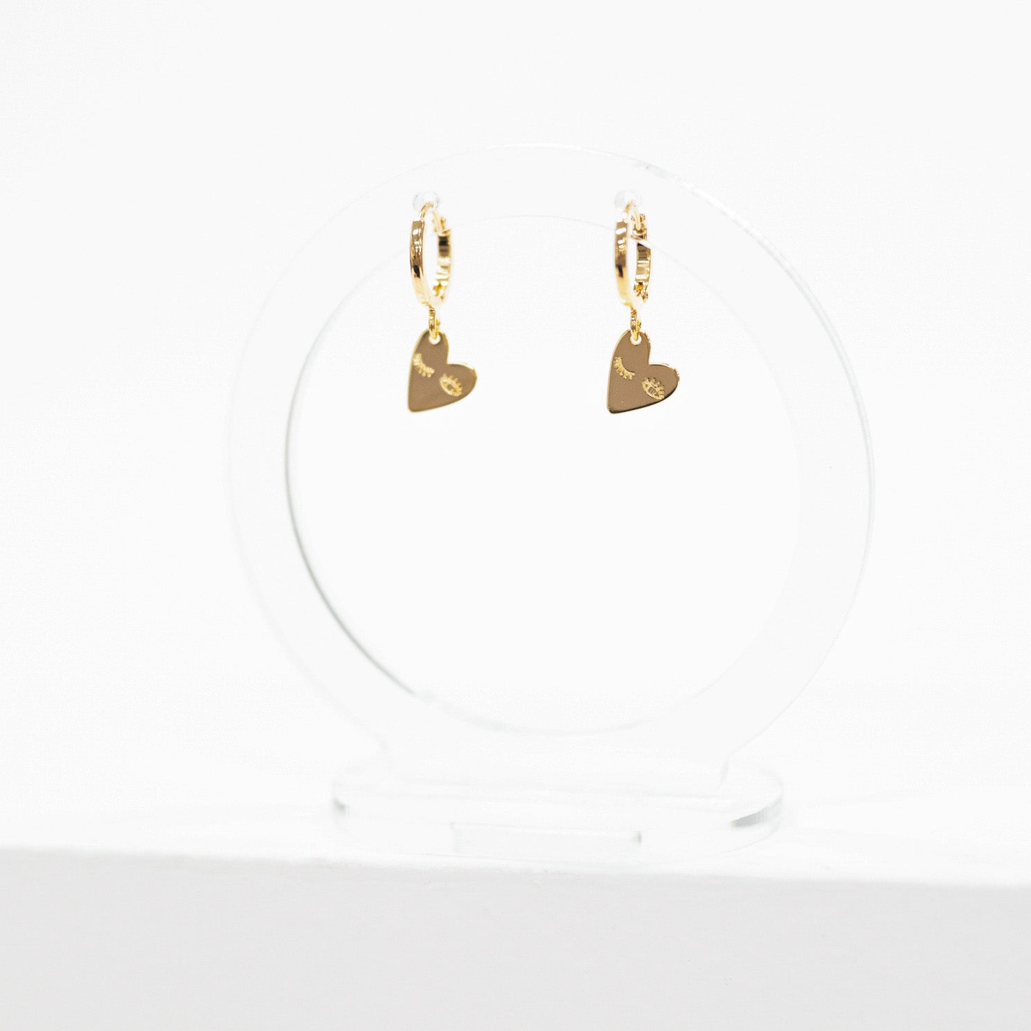 A pair of gold huggie hoop earrings with small heart charms, displayed on a clear acrylic stand against a white background.