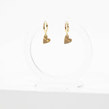 A pair of gold huggie hoop earrings with small heart charms, displayed on a clear acrylic stand against a white background.