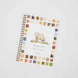 A spiral-bound watercolor workbook titled 'WOODLAND watercolor workbook' with a watercolor illustration of a bear on the cover, surrounded by a border of watercolor paint swatches, photographed on a light gray background.