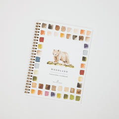 A spiral-bound watercolor workbook titled 'WOODLAND watercolor workbook' with a watercolor illustration of a bear on the cover, surrounded by a border of watercolor paint swatches, photographed on a light gray background.