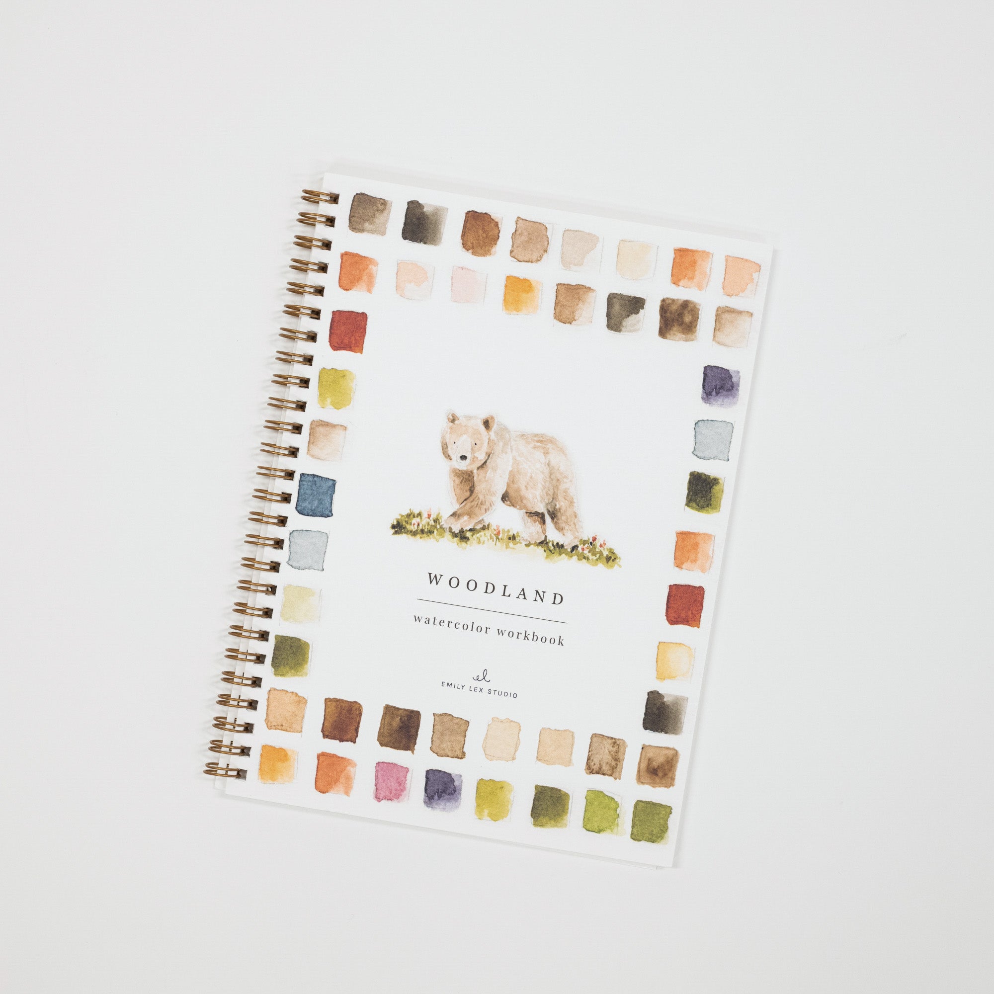 A spiral-bound watercolor workbook titled 'WOODLAND watercolor workbook' with a watercolor illustration of a bear on the cover, surrounded by a border of watercolor paint swatches, photographed on a light gray background.