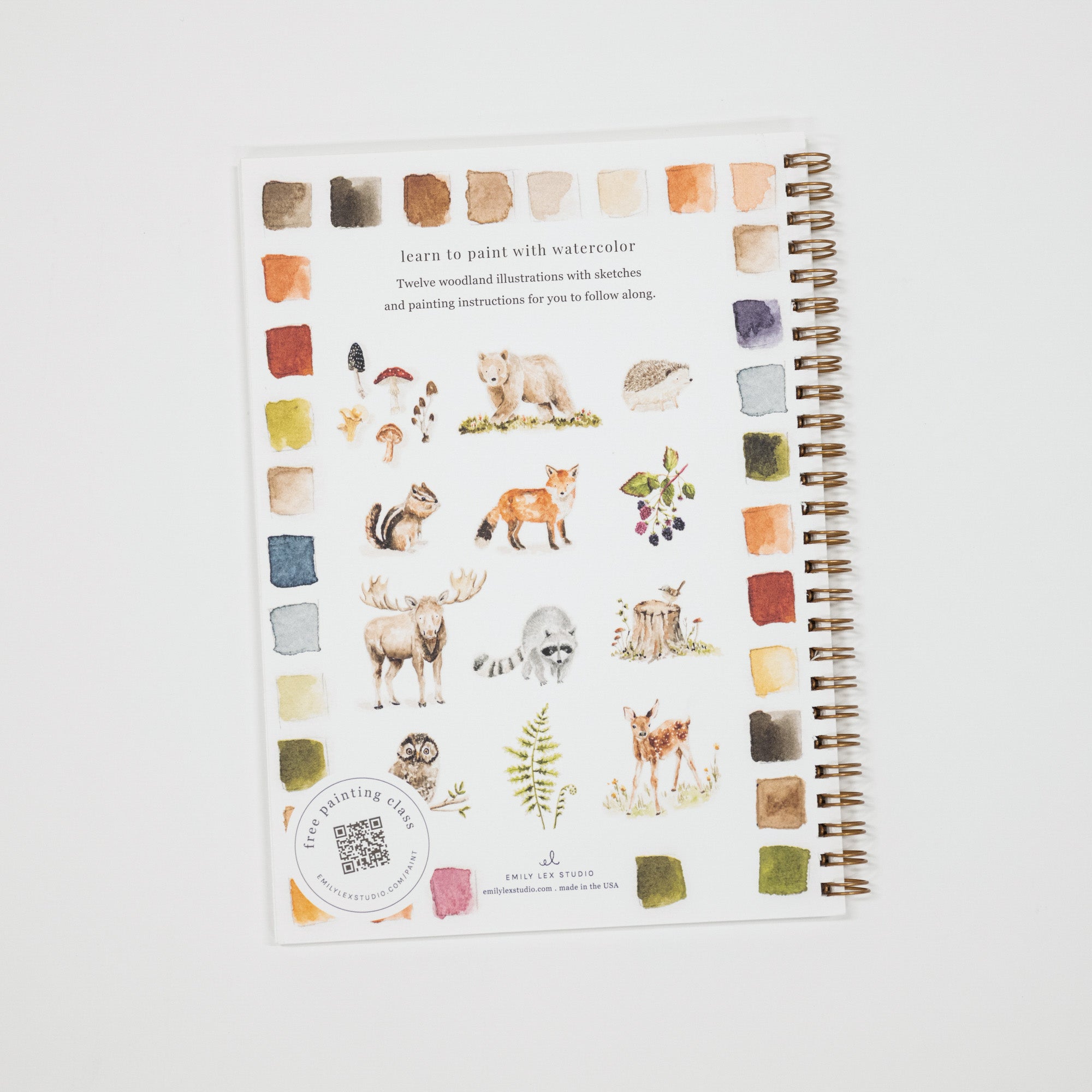A spiral-bound watercolor painting notebook with woodland illustrations of animals and trees, surrounded by watercolor swatches, photographed on a light gray background.