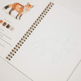 An open spiral-bound sketchbook or art journal on a white surface, showing a watercolor fox illustration with color chips and text on one page, and a faint line drawing of a fox on the next page.