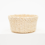 A small round woven basket made from natural beige raffia or straw fibers, photographed on a clean white surface against a white background.