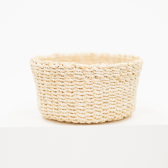 A small round woven basket made from natural beige raffia or straw fibers, photographed on a clean white surface against a white background.