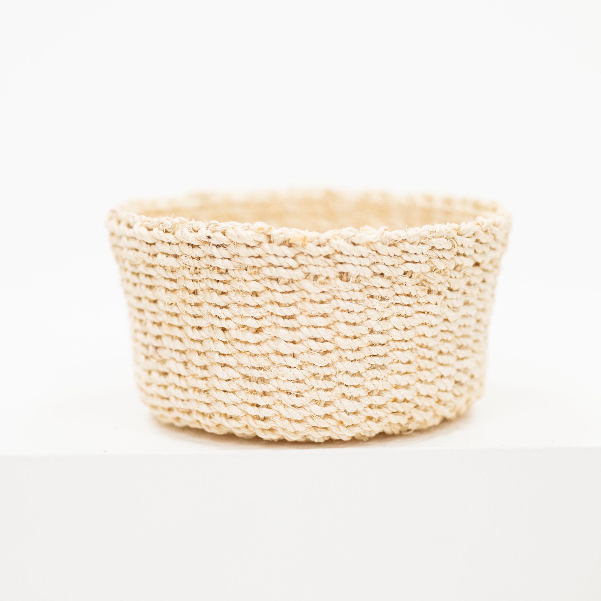 A small round woven basket made from natural beige raffia or straw fibers, photographed on a clean white surface against a white background.