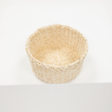 A small woven natural fiber basket in a light beige color, photographed from a top-down angle on a clean white background.