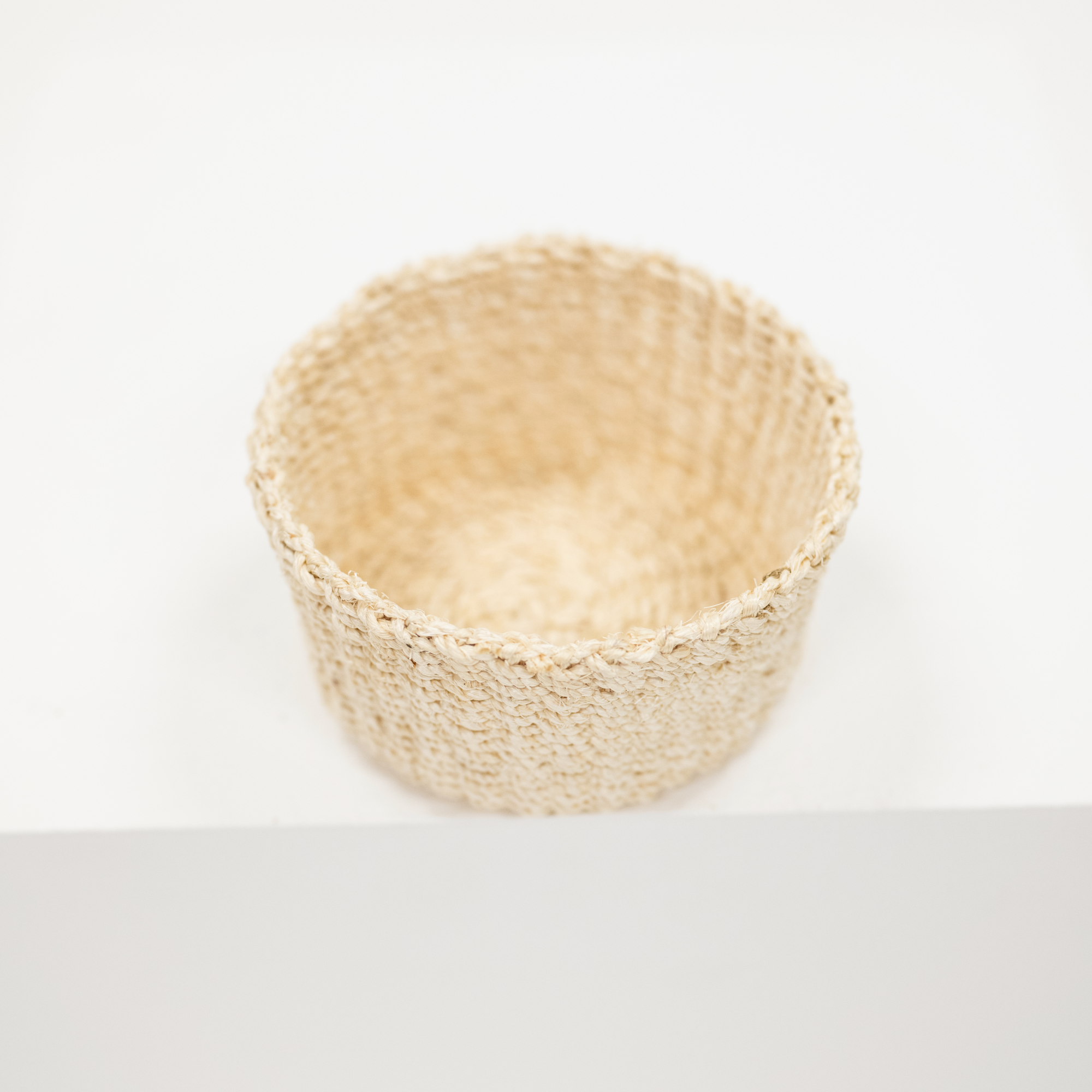 A small woven natural fiber basket in a light beige color, photographed from a top-down angle on a clean white background.